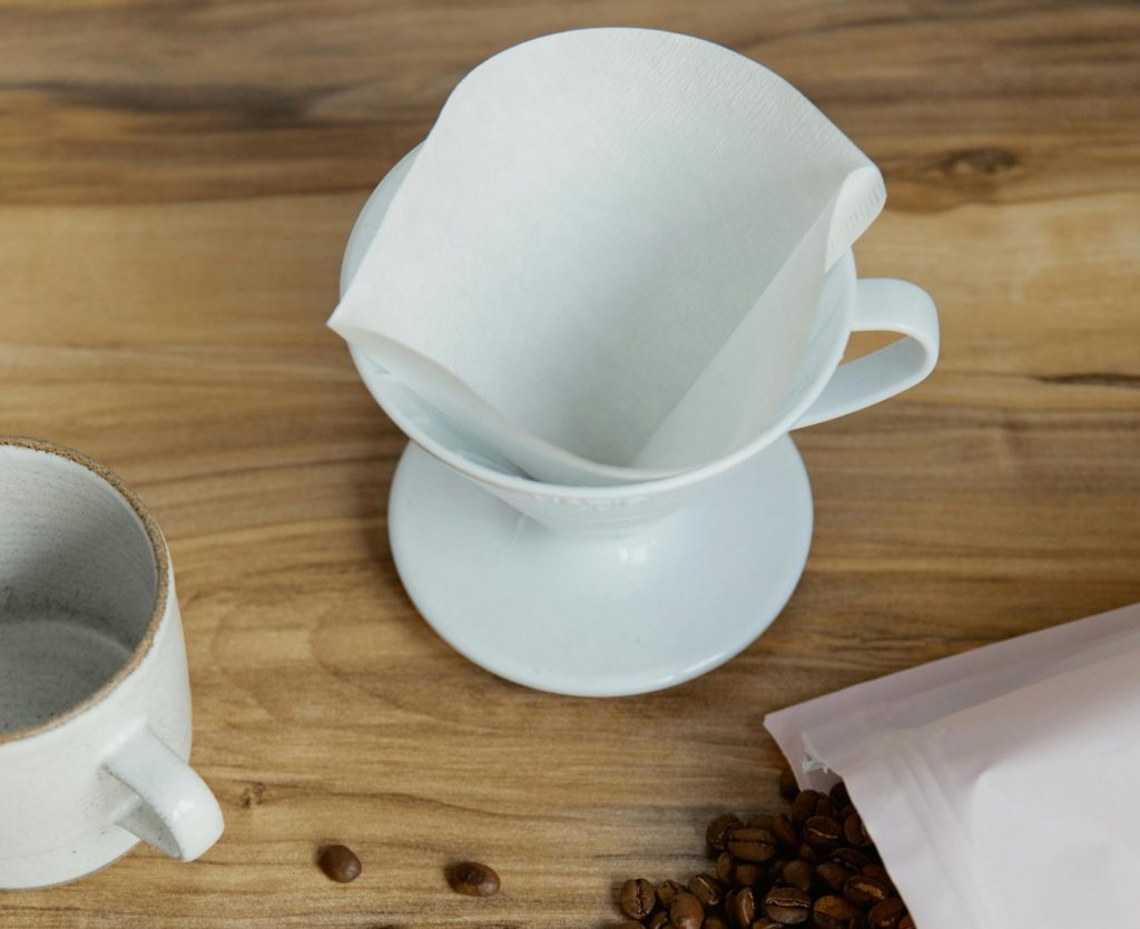 Kaffeefilter mit Filtertüte, Kaffebohnen und Kaffeetasse liegen und stehen daneben.